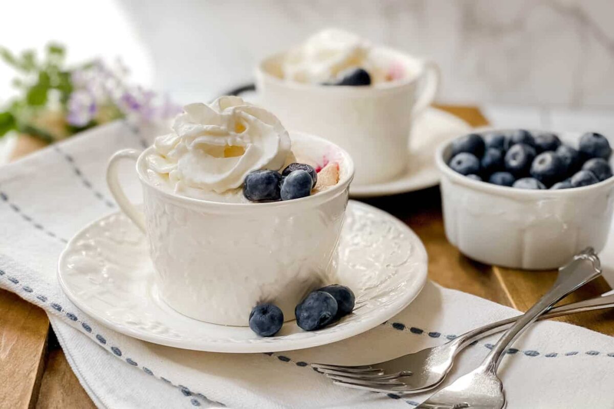Side view of 2 Blueberry Mug Cakes on a wood board with fresh blueberries