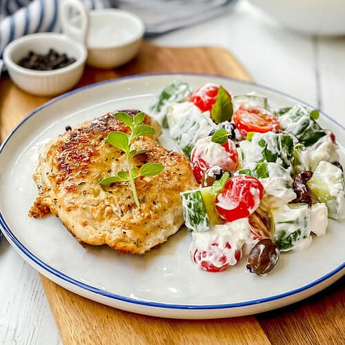 Greek Chicken with Cucumber: Grilled chicken breast garnished with herbs, served alongside a creamy salad featuring tomatoes, cucumbers, olives, and fresh greens on a white plate.