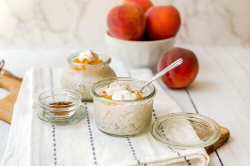 Side view of 2 Peach Overnight Oats containers with fresh peaches in the background