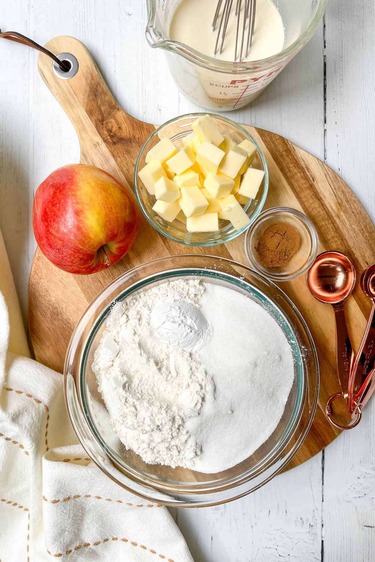 Apple Cinnamon Scones ingredients on a round wooden board to include flour, sugar, leavening, cubed butter, cream, spices, and a fresh apple.