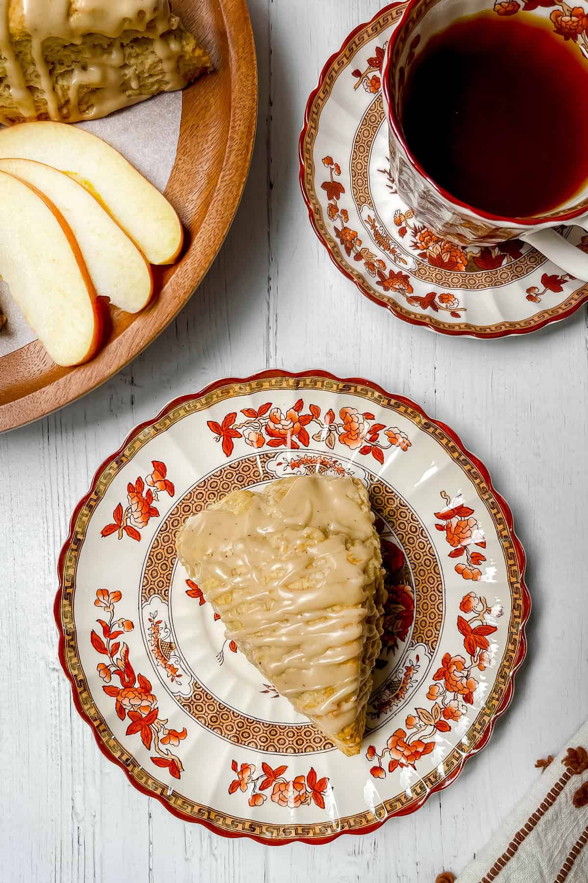 Overhead view of a glazed apple cinnamon scone on a fall patterned plate with apple slices and a cup of tea.