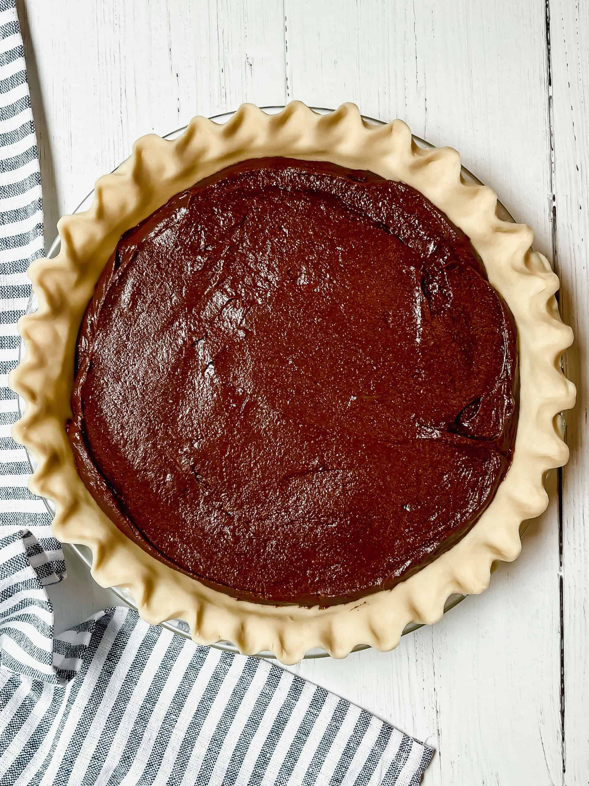 Unbaked chocolate pie filling in a fluted pie crust sits on a white wooden surface next to a striped kitchen towel, ready to become your new favorite brownie pie recipe.