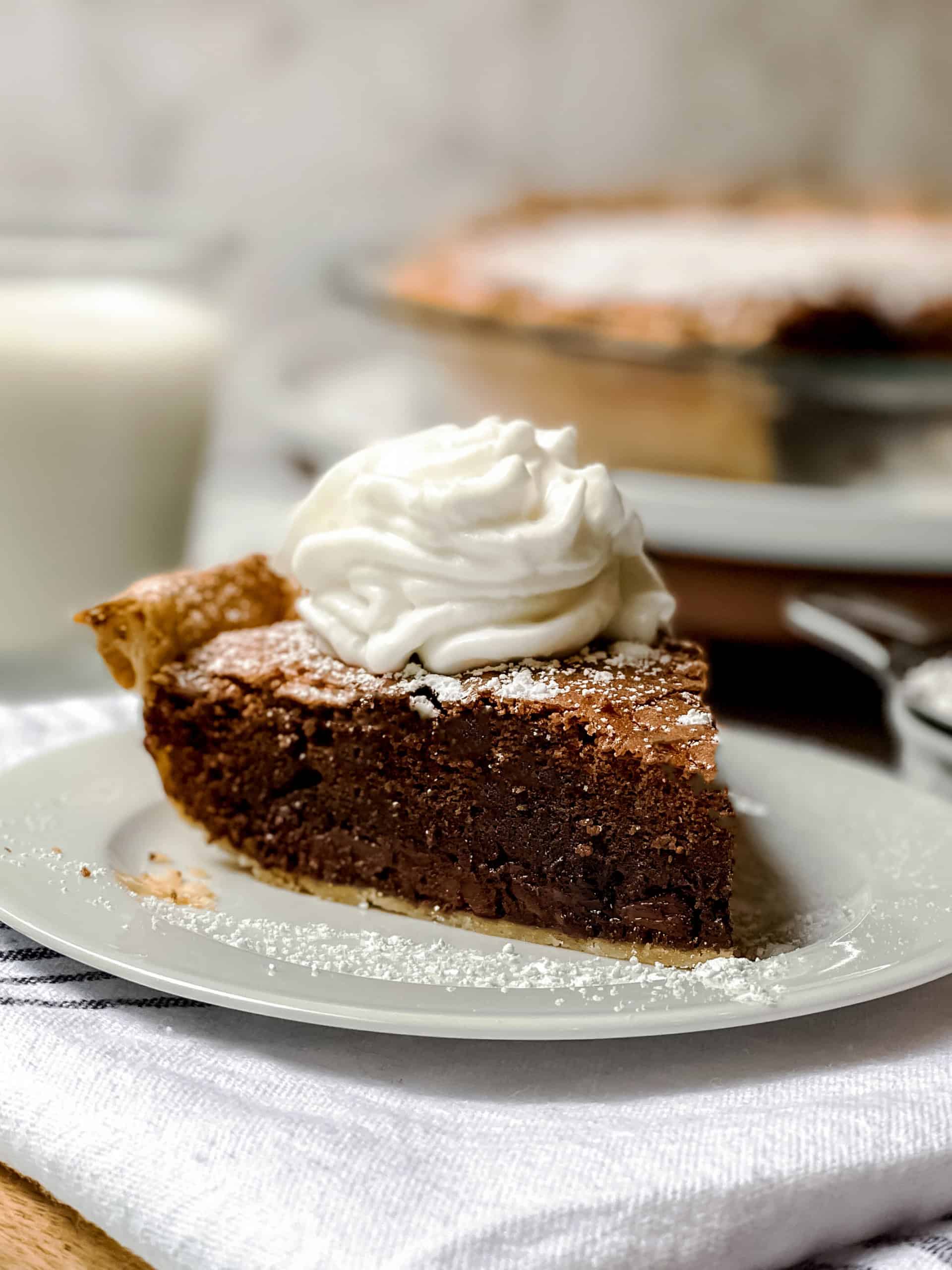 A slice of chocolate pie topped with whipped cream on a white plate, served alongside a glass of milk and a whole pie in the background&mdash;perfect inspiration for your next brownie pie recipe.
