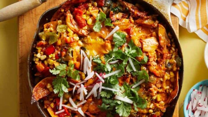 A cast iron skillet filled with a colorful Fall Mediterranean Diet chicken and vegetable casserole, topped with melted cheese, cilantro, and sliced radishes, sits on a wooden board with serving utensils nearby.