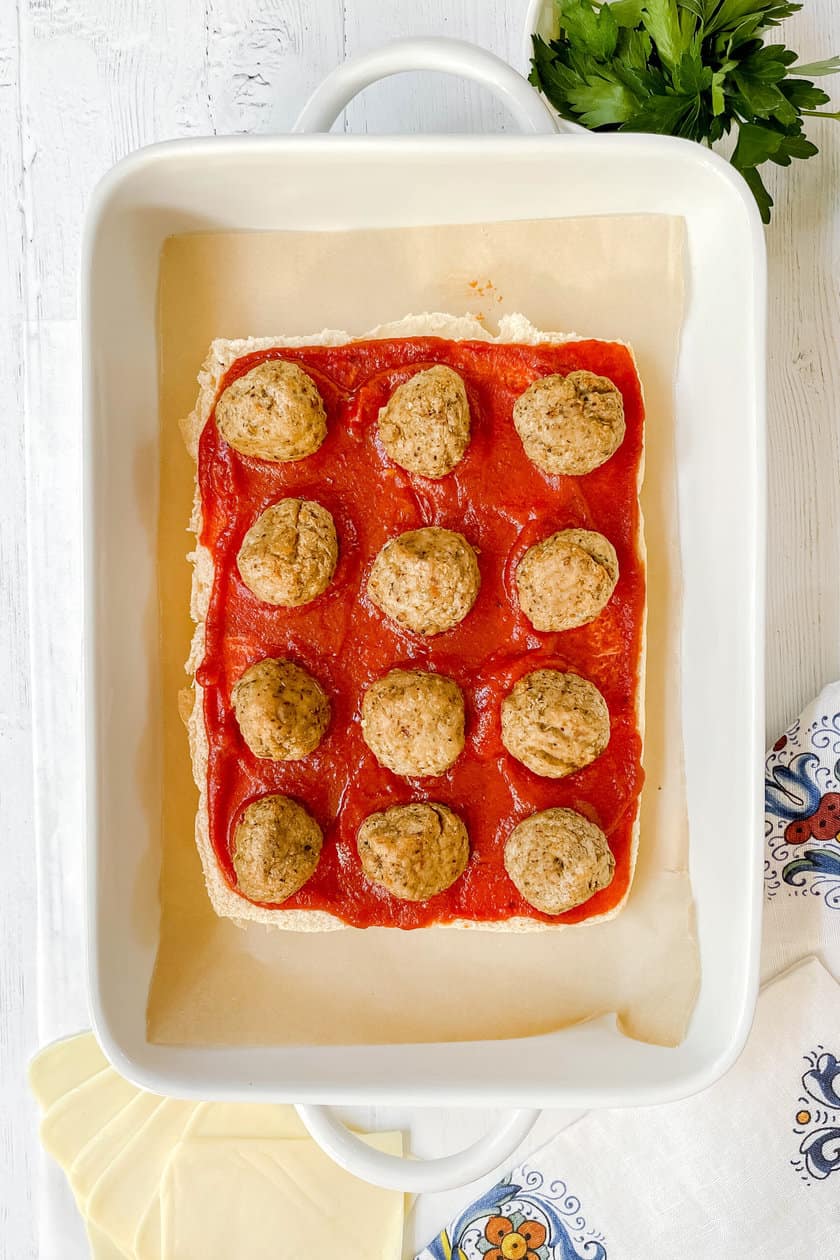 Top view of unbaked Meatball Hawaiian Roll sliders split in half and topped with marinara sauce and cooked meatballs