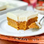 Square image of pumpkin bars with cream cheese frosting on a white plate with fork and a bite missing.