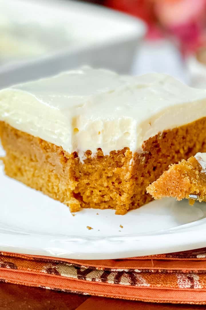 Square image of pumpkin bars with cream cheese frosting on a white plate with fork and a bite missing.