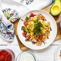 A Chicken and Rice Burrito Bowl Recipe features rice with beans, corn, tomatoes, and shredded cheese, served alongside tortilla chips, avocado, salsa, and sour cream on a white wooden table.
