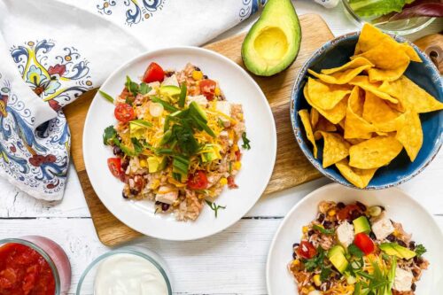 A Chicken and Rice Burrito Bowl Recipe features rice with beans, corn, tomatoes, and shredded cheese, served alongside tortilla chips, avocado, salsa, and sour cream on a white wooden table.