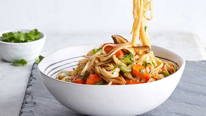 Chopsticks lift noodles with vegetables from a white bowl on a gray surface; perfect for 30 minute dinners for September, with cilantro in a small bowl in the background.