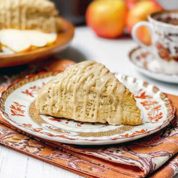 Apple cinnamon scones, drizzled with maple glaze, sits on a plate for serving with a teacup and saucer plus more scones in the background.