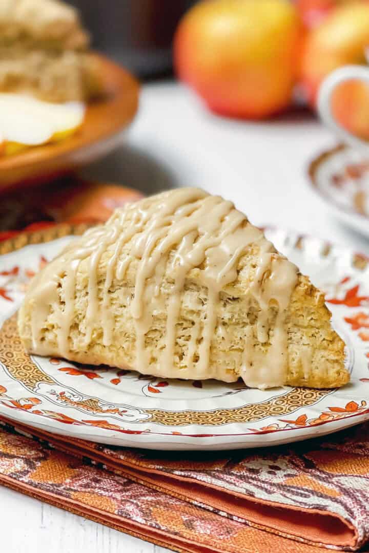 Apple cinnamon scones, drizzled with maple glaze, sits on a plate for serving with a teacup and saucer plus more scones in the background.