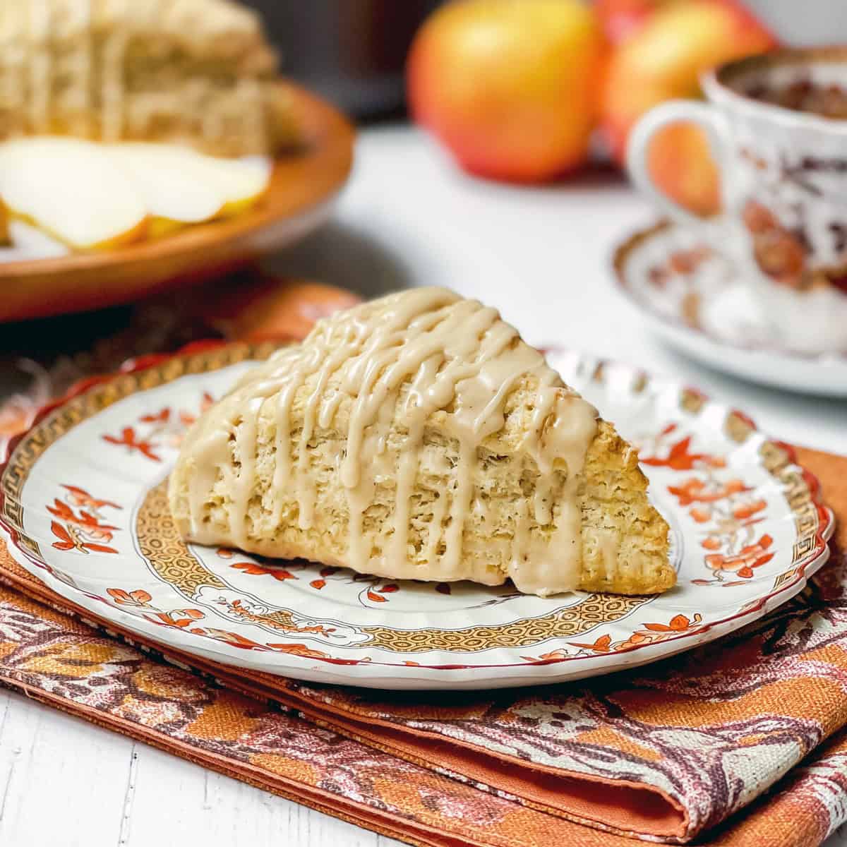 Apple cinnamon scones, drizzled with maple glaze, sits on a plate for serving with a teacup and saucer plus more scones in the background.