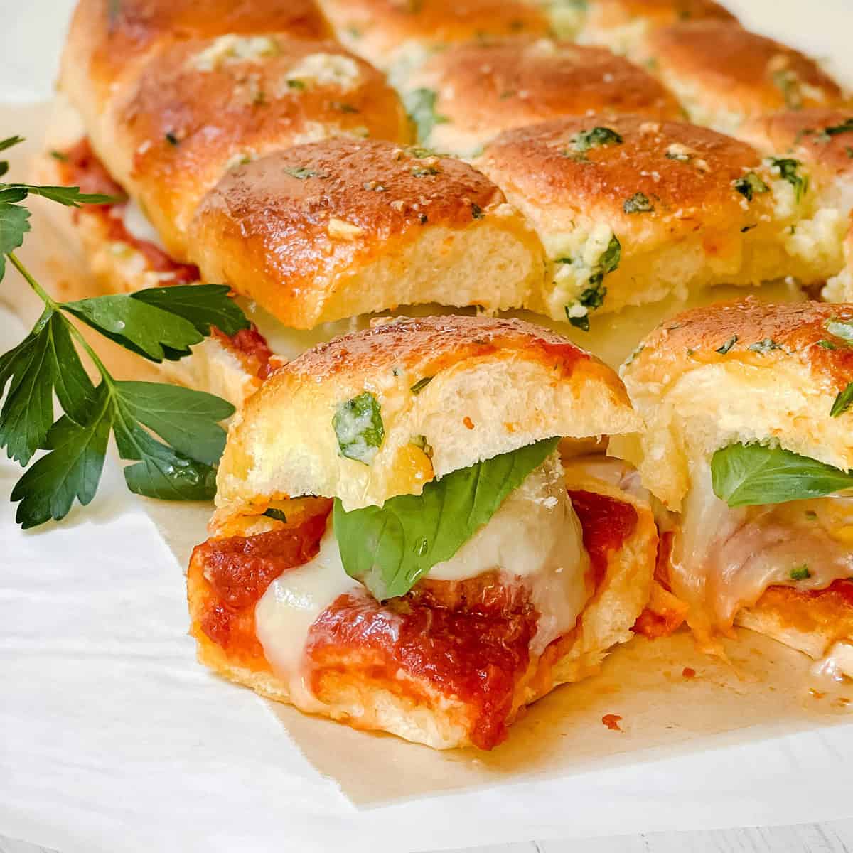 Close-up of Hawaiian Roll Sliders: pull-apart bread sliders filled with melted cheese, tomato sauce, and basil, topped with a golden, herbed crust and garnished with parsley.