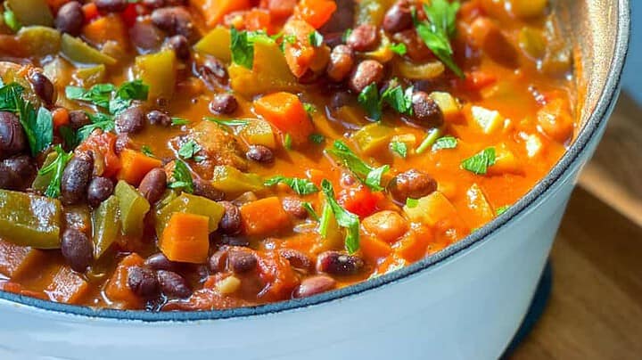 A white pot filled with vegetarian chili made of vegetables and black beans, topped with chopped herbs, sits on a wooden surface with cornbread in the background.