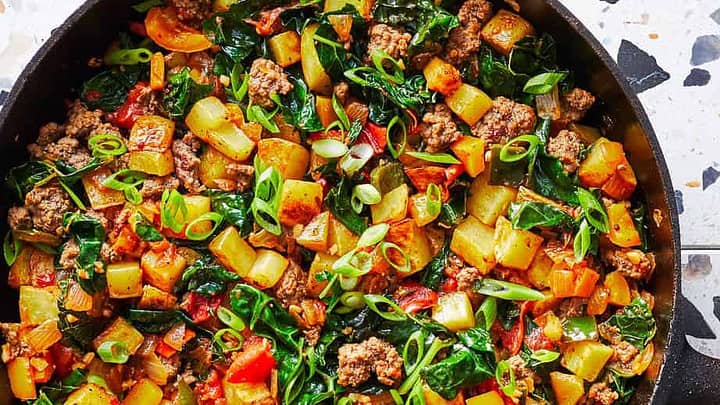 A skillet filled with a cooked mixture of ground meat, diced potatoes, spinach, tomatoes, and green onions makes for hearty October Skillet Dinners, with a wooden spoon resting inside.