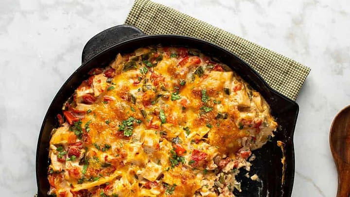 A cast iron skillet filled with a baked cheesy casserole of chopped vegetables and herbs, perfect for October 30 Minute Dinners, placed on a cloth napkin next to a wooden spoon.