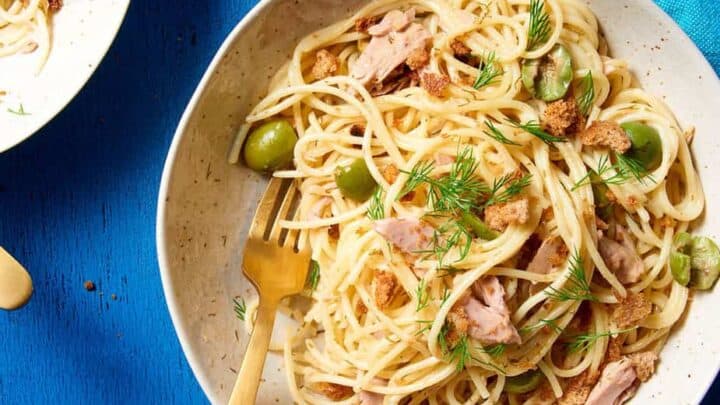 A bowl of spaghetti with tuna, green olives, fresh dill, and breadcrumbs—an ideal October One Pot Dinner—served with a gold fork on a blue table.