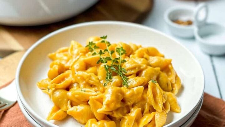 A plate of creamy pumpkin mac and cheese garnished with fresh herbs sits on a cloth napkin, with a pot of more pasta and small pumpkins in the background.