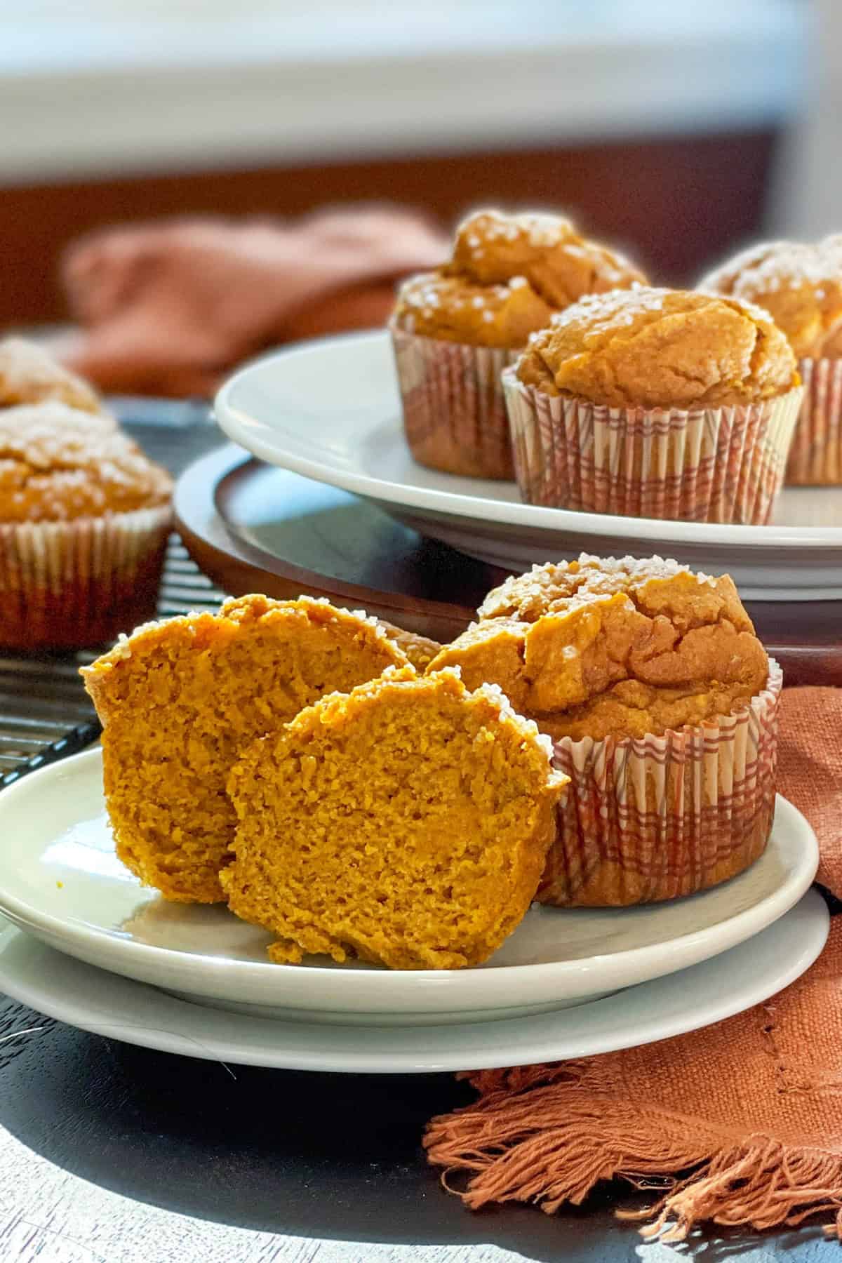 A plate with two healthy pumpkin muffins, one sliced in half, sits on a table with more muffins on a cooling rack and a white plate in the background.