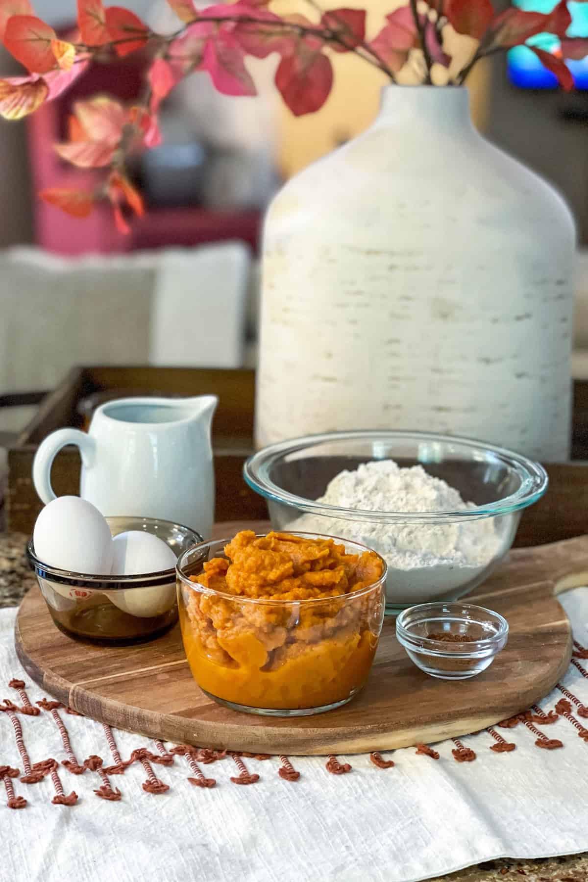 A wooden board holds ingredients for healthy pumpkin muffins: two eggs, a glass bowl of mashed sweet potatoes, a bowl of flour, a small bowl of clear liquid, and a small pitcher of milk on the countertop.