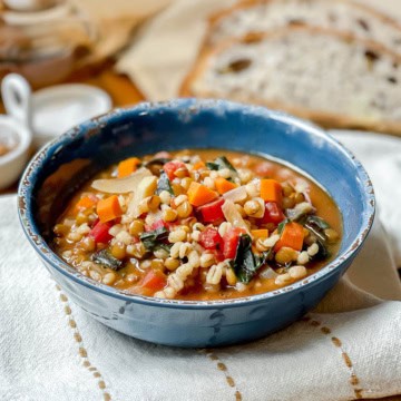 A blue bowl filled with vegetable and barley soup, inspired by a classic lentil soup recipe, features carrots, greens, and tomatoes. It sits on a white cloth with slices of bread in the background.