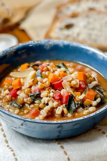 A blue bowl filled with vegetable and barley soup, inspired by a classic lentil soup recipe, features carrots, greens, and tomatoes. It sits on a white cloth with slices of bread in the background.