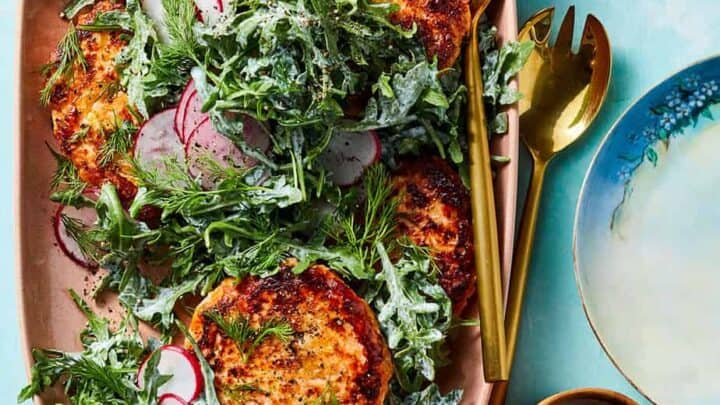 A platter with salmon cakes topped with arugula, sliced radishes, and dill—perfect for November healthy dinners—served with gold utensils, a sauce cup, and a decorative plate on a light blue surface.