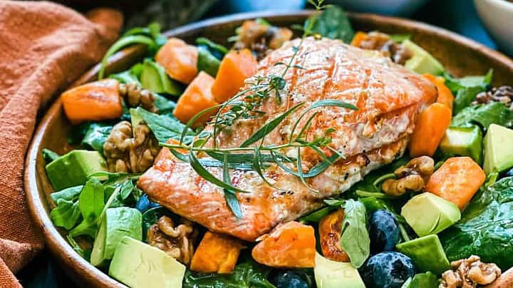 A vibrant salad featuring a grilled salmon fillet garnished with herbs. The salad includes fresh spinach, avocado chunks, blueberries, diced sweet potatoes, and walnuts. Accompanying the dish are small bowls containing additional sweet potatoes and walnuts.