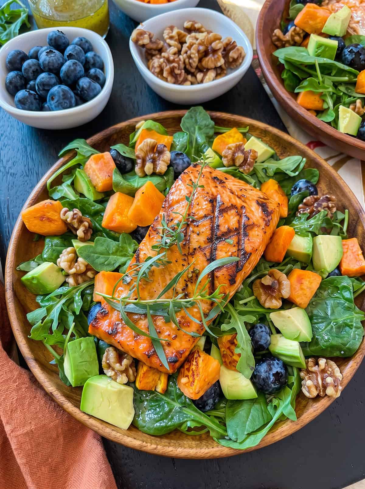 A wooden bowl filled with a vibrant salmon salad-salad greens, cooked salmon fillet, roasted sweet potatoes, blueberries, walnuts, and avocado.