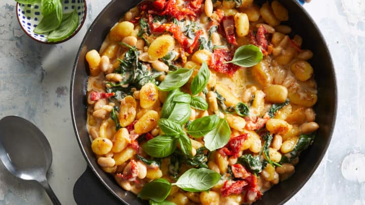 A skillet filled with gnocchi, spinach, white beans, sun-dried tomatoes, and melted cheese, garnished with fresh basil leaves—an ideal choice for October 30 Minute Dinners. A spoon and a small bowl of basil sit beside the skillet.