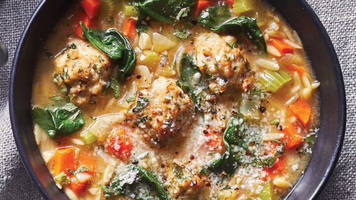 A bowl of soup with meatballs, orzo pasta, spinach, carrots, celery, and herbs, garnished with grated cheese—perfect for quick November 20 Minute Dinners—served on a gray fabric surface.