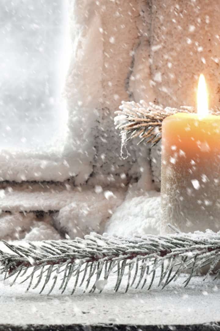 A lit candle sits on a snowy windowsill with evergreen branches, making Christmas memorable as snow falls outside a frosted window.