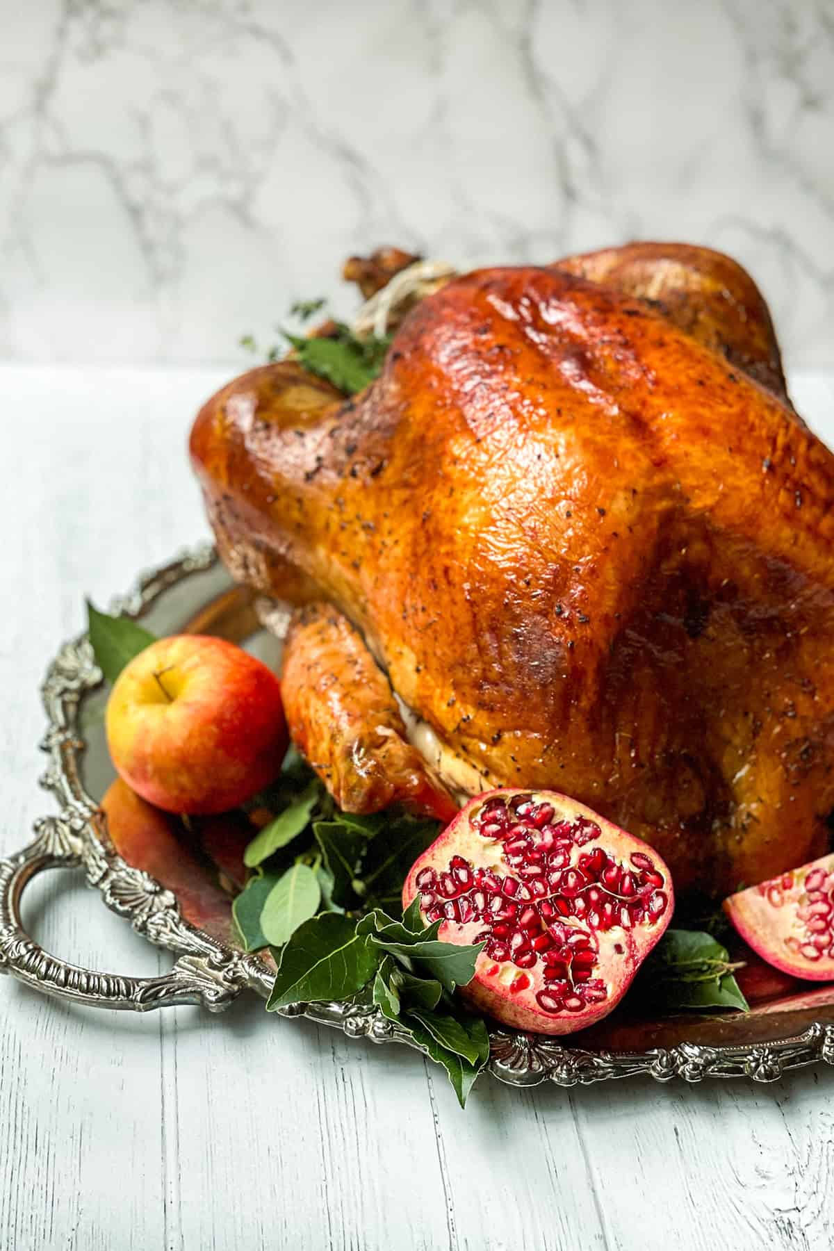Whole herb roasted roast turkey on a silver platter with bay leaves, apples, and pomegranates as garnish.