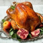 Square image of herb roast turkey on a silver platter with apples, bay leaves, and cut pomegranates.