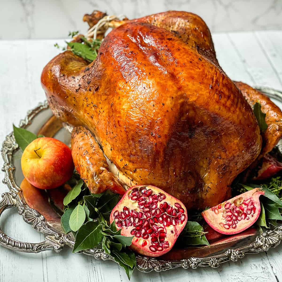 Square image of herb roast turkey on a silver platter with apples, bay leaves, and cut pomegranates.