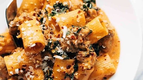 A white plate of rigatoni pasta in a creamy tomato sauce with sausage, kale, and grated cheese&mdash;perfect for cozy December dinners&mdash;sits next to a fork and a dark napkin.