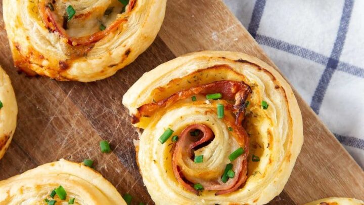 Ham and cheese puff pastry appetizers are served as pinwheels on a wooden board, garnished with chopped chives, with a small bowl of chives in the background.