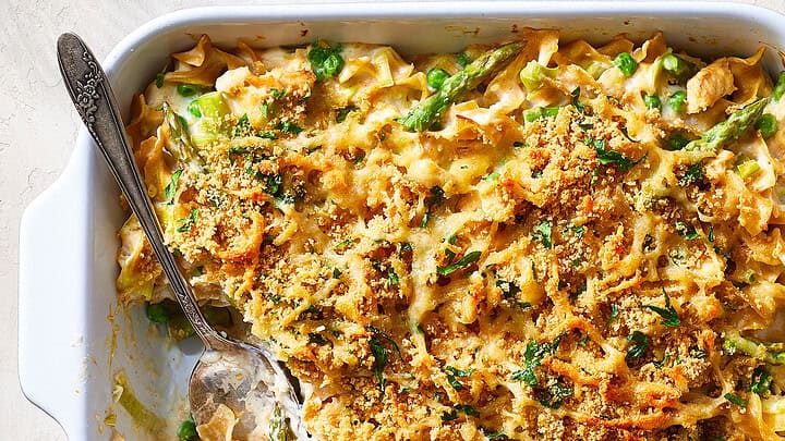 A baked casserole dish filled with noodles, peas, and a creamy sauce, topped with golden breadcrumbs and herbs—perfect comfort food for healthy December dinners—with a serving spoon partially removing a portion.