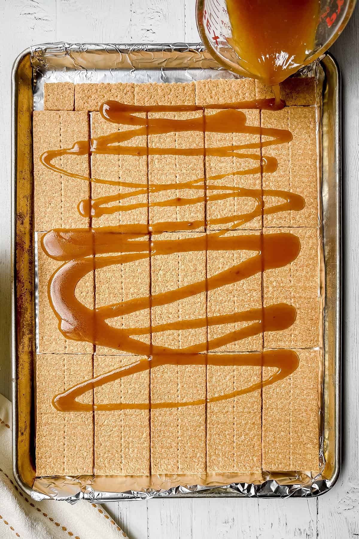 Graham crackers fitted into a foil lined baking sheet with hot toffee being poured over the top.