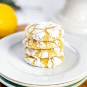 A stack of 4 lemon crinkle cookies, dusted with powdered sugar, on a white plate with fresh lemons in the background.