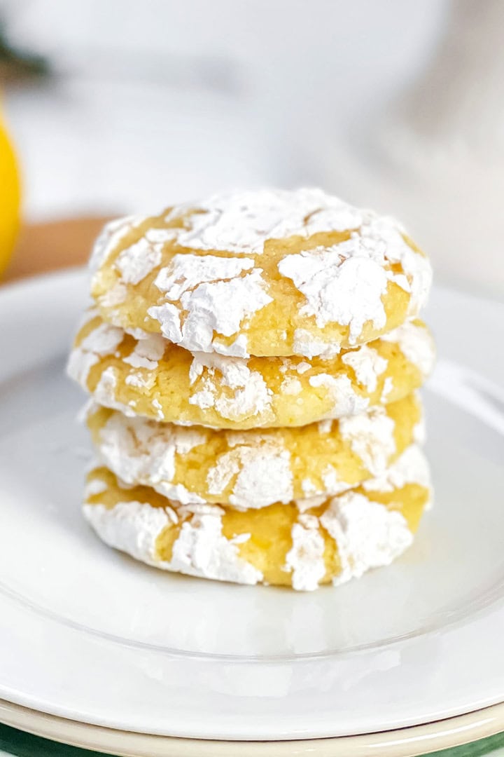 A stack of 4 lemon crinkle cookies, dusted with powdered sugar, on a white plate with fresh lemons in the background.