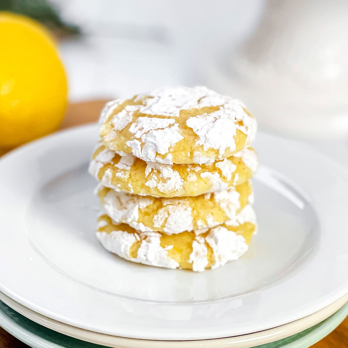 A stack of 4 lemon crinkle cookies, dusted with powdered sugar, on a white plate with fresh lemons in the background.