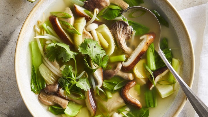 A bowl of clear soup with assorted mushrooms, bok choy, green onions, cilantro, and a wedge of lime on the side—perfect for Healthy Winter Dinners. A spoon is placed in the bowl.
