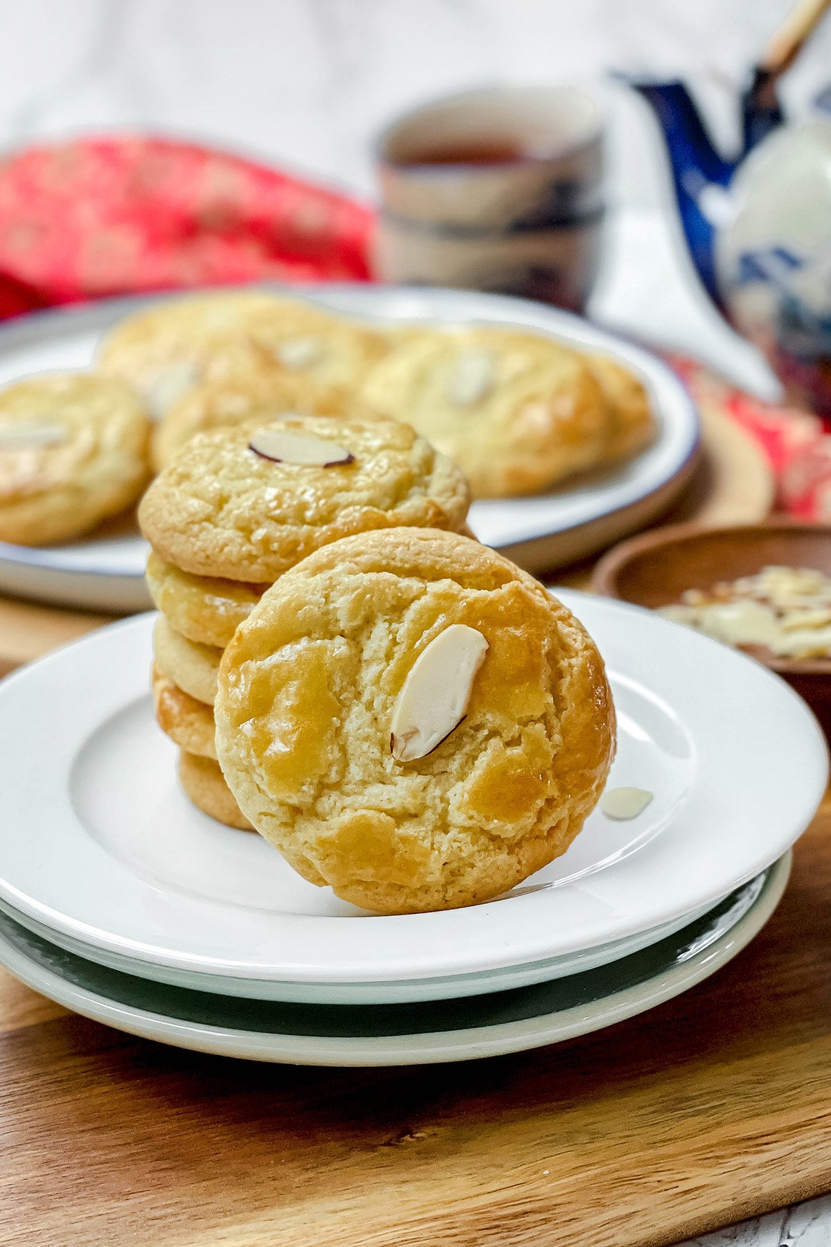 A stack of Chinese Almond Cookies is displayed on white plates, with more cookies and a teapot set in the background.