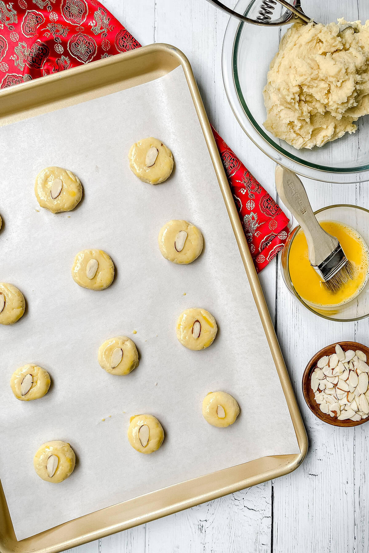 A baking sheet with unbaked Chinese Almond Cookies topped with almond slices on parchment paper, next to a bowl of dough, egg wash, almond slices, and a pastry brush.