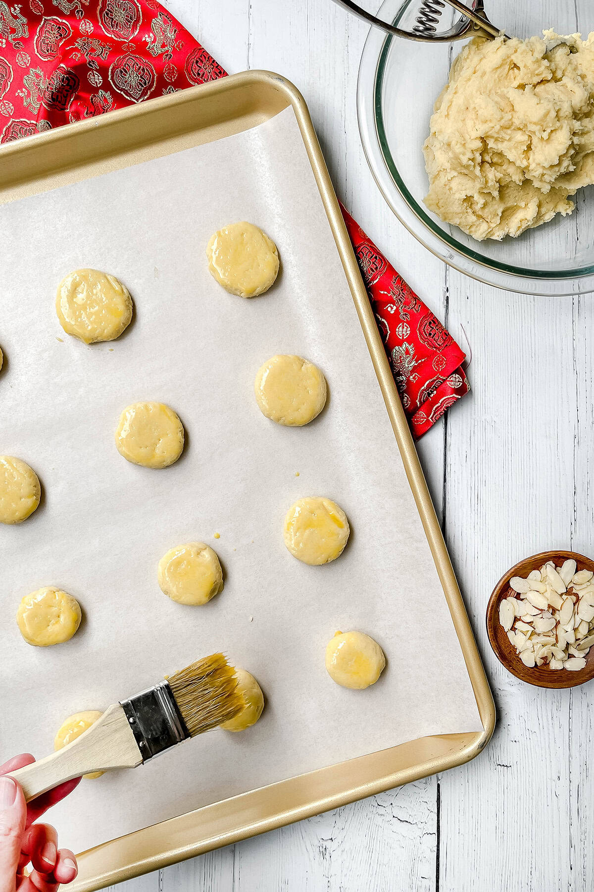 A hand brushes egg wash onto round Chinese Almond Cookies dough pieces on a parchment-lined baking sheet; a bowl of dough and sliced almonds are nearby.