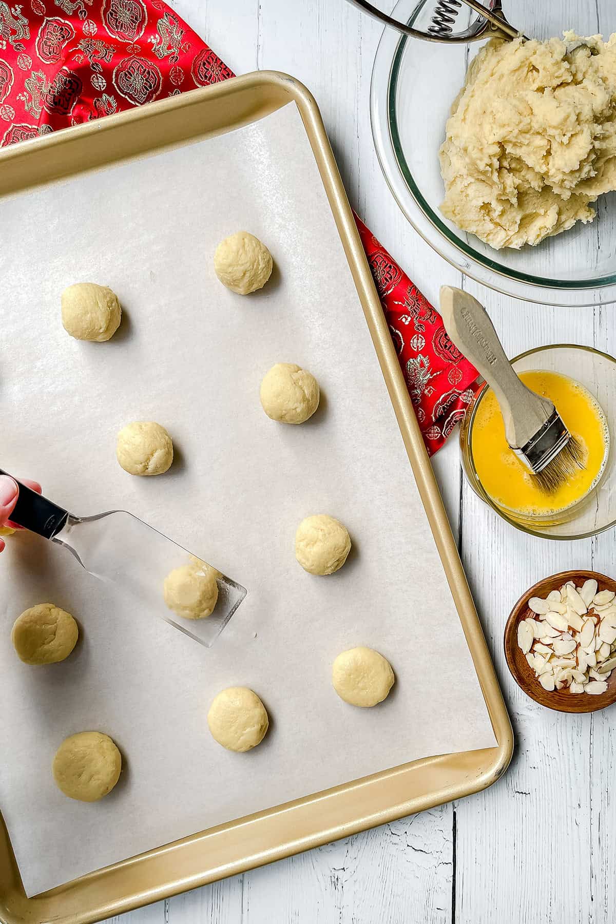 Unbaked Chinese Almond Cookies dough balls arranged on a parchment-lined baking sheet with a scoop, next to bowls of beaten egg, sliced almonds, and more dough on a white wooden surface.