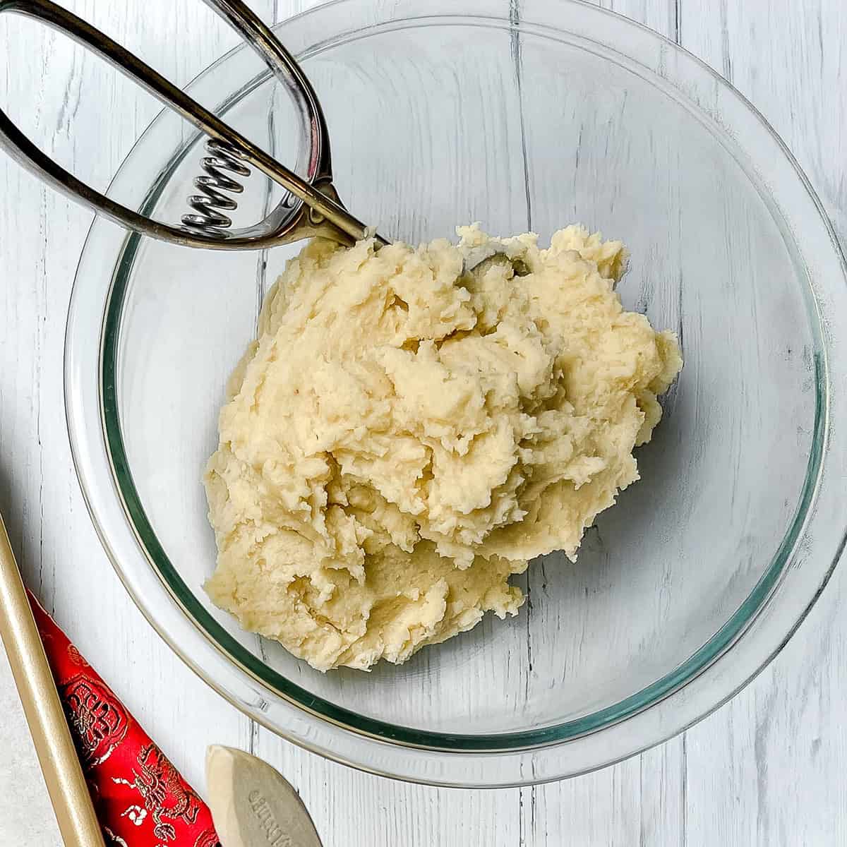 A glass bowl containing Chinese Almond Cookies dough with a stainless steel scoop resting inside, placed on a white wooden surface.