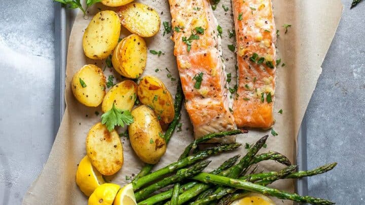 Baked salmon fillets, roasted baby potatoes, and asparagus spears garnished with parsley and lemon wedges on a parchment-lined baking sheet make for delicious anti inflammatory dinners.